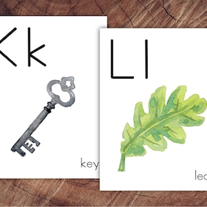 May include: Two white cards with the letters "Kk" and "Ll". The "Kk" card features a gray key and the word "key". The "Ll" card shows a green leaf and the word "leaf". The cards are on a wooden surface.