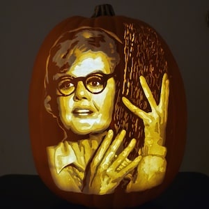May include: A carved pumpkin with a detailed image of a woman with glasses and a hand reaching out. The pumpkin is lit from inside, creating a warm glow.
