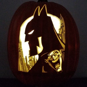 May include: A carved pumpkin with a silhouette of Batman and the Joker. The pumpkin is lit from inside, creating a glow effect. The Joker is grinning with a wide, toothy smile.