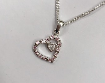 Peut inclure: Un collier en argent avec un pendentif en forme de cœur. Le pendentif a un plus petit cœur à l'intérieur, tous deux sertis de pierres roses et claires.