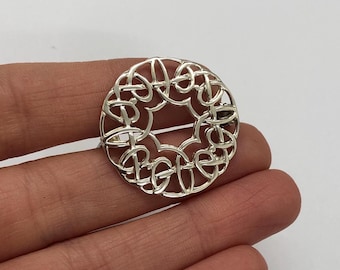 Pode incluir: Broche de prata com design circular de nós celtas. O design intrincado apresenta um padrão repetitivo de nós entrelaçados.