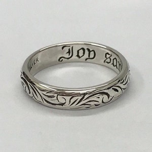 May include: A silver band ring with an intricate floral design and the words "Joy" and "Save" engraved on the inside.