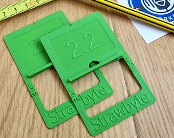 Op de afbeelding: Twee groene kunststof standaards met het nummer "2.2" erop gedrukt. De standaards zijn gelabeld met "Strawbyte" en hebben een klein gleufje voor een telefoon of tablet.
