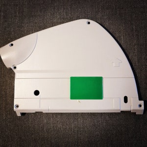 Puede incluir: Una pieza de plástico blanca con una pegatina rectangular verde. La pieza tiene un pequeño agujero y un símbolo de flecha en relieve.