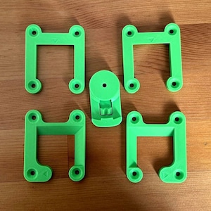 May include: Four bright green, U-shaped mounting brackets with four circular screw holes each, arranged around a central circular component featuring a small lever mechanism and a single hole in the middle, all with a matte, textured finish.