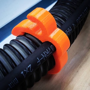 Puede incluir: Abrazadera de cable de plástico naranja que sujeta un cable negro y una tubería corrugada.