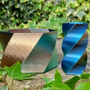 May include: Two decorative geometric vases. One is a square shape with a gradient of brown, gold, and green. The other is a tall, twisted vase with a blue and teal gradient. Both are on a stone surface with green foliage.