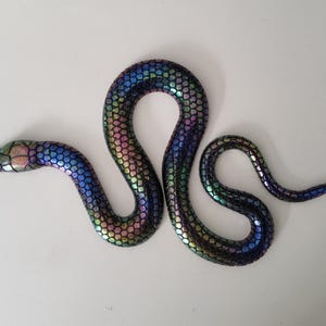 Könnte beinhalten: Dekorative Schlangenfigur in schillernden Farben von Blau, Grün, Rosa und Lila. Die Schlange hat ein strukturiertes, schuppenartiges Muster und ist gewunden. Der Kopf ist leicht angehoben, und der Körper ist anmutig geschwungen.