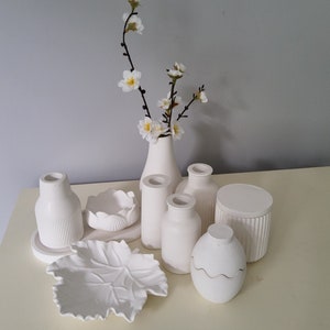 Pode incluir: Um conjunto de vasos, tigelas e recipientes de cerâmica branca, incluindo um prato em forma de folha, uma tigela pequena com borda canelada e um vaso com um arranjo floral delicado. Os itens estão dispostos em uma superfície branca.
