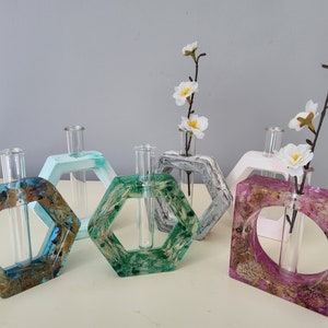 Puede incluir: Cinco jarrones de flores hexagonales y cuadrados de resina con tubos de ensayo de vidrio. Los jarrones son de varios colores, incluyendo turquesa, verde, gris, blanco y rosa. Cada jarrón tiene flores secas incrustadas en la resina.