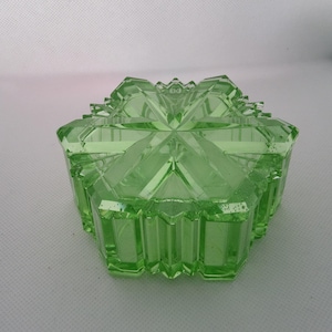 Pode incluir: Uma caixa de bugigangas hexagonal de vidro verde claro com um design geométrico. A caixa tem uma tampa com um padrão de estrela e uma borda facetada. A caixa é feita de um material de vidro translúcido.