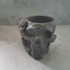 May include: A grey skull-shaped planter with a detailed tiger design on the side. The planter has a large opening for plants and a textured surface. The tiger has yellow eyes and is in shades of brown and grey.