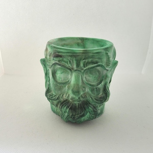May include: A green ceramic planter in the shape of a man's face with a beard and glasses. The planter has a textured finish and a hollow interior for plants or other items. The colour is a mottled green with darker accents.