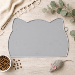 May include: A light gray silicone pet feeding mat shaped like a cat head with embossed ear, whisker, and eye details. The mat features the text LINDO LEO embossed at the bottom center and includes a small heart-shaped cutout on the right side edge.