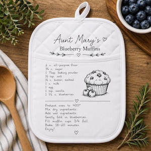 May include: A white quilted pot holder featuring a printed recipe for Aunt Mary's Blueberry Muffins. The design includes a list of ingredients, a line drawing of a muffin with blueberries, and step-by-step baking instructions in black text.