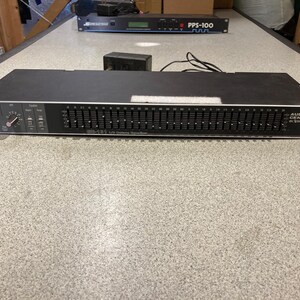 May include: A black Audio Centron EQ-131 1/3 octave equalizer rack-mount unit featuring a series of vertical sliders for frequency adjustment, a level control knob, and input/output jacks on the front panel, with a power cord attached at the rear.