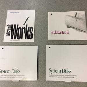May include: Four vintage Apple computer manuals and software disks are arranged on a gray surface. The manuals include "Microsoft Works" and "StyleWriter II." Two packages are labeled "System Disks."