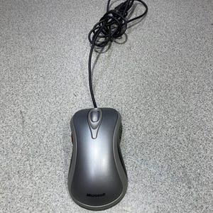 May include: A wired Microsoft computer mouse featuring a smooth, dark gray contoured shell with a lighter gray border. The device includes a central scroll wheel and a black cord that is coiled above the main body of the mouse.
