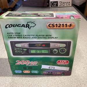 May include: A Cougar CS1211-F car stereo cassette player box with a green and white gradient design. The packaging displays the text Auto Stop, AM/FM-MPX Radio, Digital Display, 200W P.M.P.O., and LCD Display. A white sticker with the number 473 is attached.