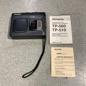 May include: A dark gray Aiwa cassette recorder with a black wrist strap, accompanied by an operating instructions manual for models TP-560 and TP-510, and a printed Aiwa America Inc. limited warranty document with contact information.