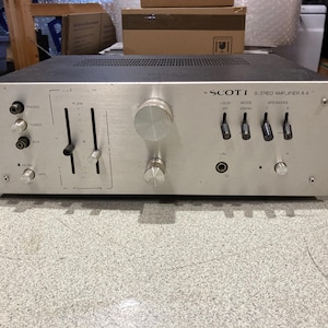 May include: A silver Scott Stereo Amplifier A 4 with a black top casing. The front panel features two vertical sliders, two large circular knobs, four toggle switches, a headphone jack, and several input selection buttons labeled Phono, Tuner, and Aux.