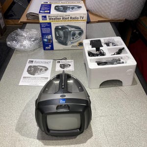 May include: A Stormtracker Series Vector Weather Alert Radio-TV with a gray and black plastic housing, a top handle, and a small screen. The set includes the original cardboard box, a white styrofoam insert, power cables, and a user manual.