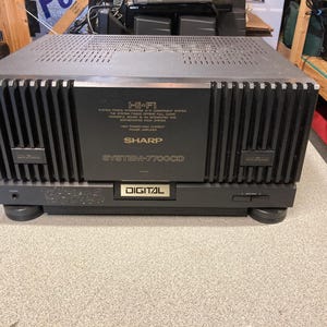 May include: A black Sharp System-7700CD Hi-Fi power amplifier featuring a central panel with gold text, vertical cooling fins on the sides, and a digital display label. The unit has a vented top cover, a front headphone jack, and four circular feet.