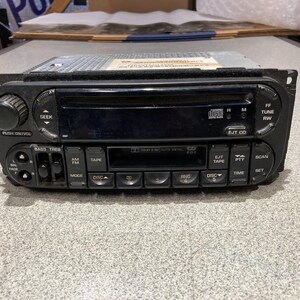 May include: A black automotive head unit featuring a CD player slot, a cassette tape deck, and a digital display. The faceplate includes a volume knob, seek buttons, tuning controls, and various function buttons labeled AM/FM, TAPE, SCAN, and TIME.