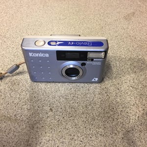 May include: A silver Konica Revio-CL digital camera with a blue accent on top. The camera has a lens, flash, and a wrist strap. The camera is on a textured surface.