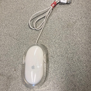 May include: A white Apple computer mouse with a clear plastic outer shell and a white USB cable. The cable is secured with a small red rubber band and features a white USB connector with an Apple logo printed on the top surface of the plug.