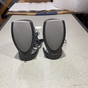 May include: A pair of Harman Kardon computer speakers with a black frame and a light gray mesh grille. The left speaker features a circular volume control knob on the front base, while the right speaker displays the Harman Kardon brand logo.