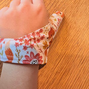 Puede incluir: Una férula para pulgar de tela blanca con un patrón repetitivo de zorros naranjas, flores rojas y follaje verde. La férula envuelve la base del pulgar y se asegura alrededor de la muñeca con costuras visibles a lo largo de los bordes.