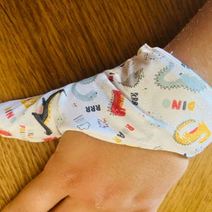May include: A white fabric finger puppet with a dinosaur pattern. The puppet features colorful dinosaur illustrations in red, yellow, blue, and gray, with the word "DINO" printed on it. The puppet is on a wooden surface.