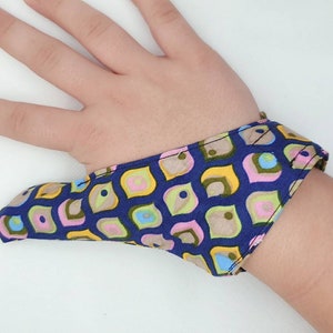 Peut inclure: Un support de pouce bleu avec un motif géométrique de formes jaunes, roses et vertes. Le support de pouce est en tissu et se porte sur le pouce et la main.