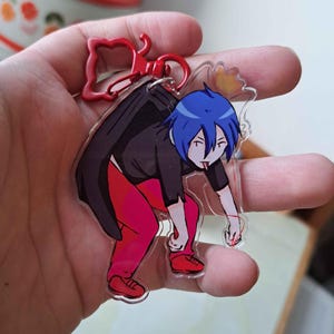 Puede incluir: Un llavero acrílico transparente con un personaje de dibujos animados con cabello azul, una camisa negra, pantalones rojos y zapatos rojos. El llavero tiene un cierre rojo y un pequeño dije. El personaje está sacando la lengua.