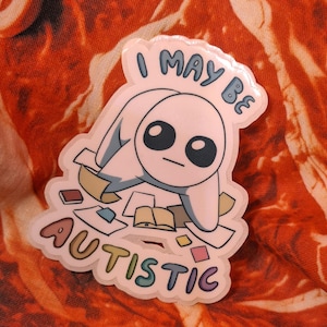 May include: A white and blue cartoon character pin with the text "I MAY BE AUTISTIC". The pin features a character surrounded by books and papers. The text is in a rainbow color scheme.