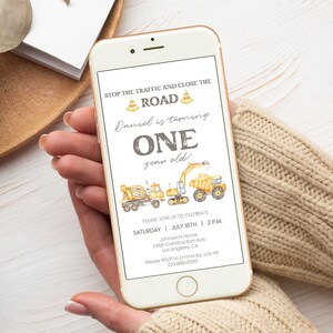 May include: A smartphone displays a construction-themed birthday invitation. The invitation features the text "STOP THE TRAFFIC AND CLOSE THE ROAD" and "ONE year old!" with illustrations of construction vehicles. The phone is held by a person.
