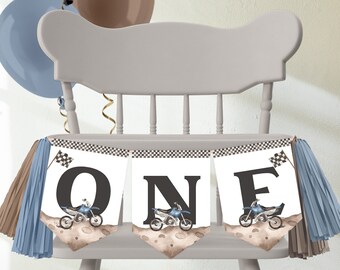 Puede incluir: Una silla de madera gris decorada con una pancarta que dice "ONE" con ilustraciones de motocicletas. La pancarta tiene borlas azules y marrones a cada lado. Tres globos en negro, marrón y azul están atados a la silla.