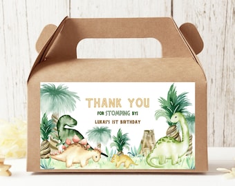 May include: A brown cardboard favor box with a handle, featuring a dinosaur-themed label. The label includes the text "THANK YOU FOR STOMPING BY! LUKAS'S 1ST BIRTHDAY" with illustrations of dinosaurs and palm trees.