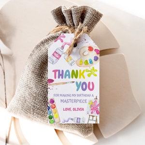 May include: A white thank you tag with colorful paint splatters and art supplies. The text reads "Thank You" in rainbow letters and "For making my birthday a masterpiece. Love, Olivia."
