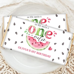 以下が含まれることがあります： 白い包装紙のチョコレートバー2本。スイカをテーマにしたデザインで、スイカのスライス、「one in a melon」、そして「Olivia's 1st Birthday」の文字が描かれています。黒い種子のイラストが包装紙全体に散りばめられています。