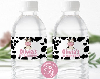 May include: Two clear plastic water bottles with black and white cow print labels. The labels have a cartoon cow with pink ears and nose and the text "Olivia's 1st Birthday".