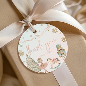 Puede incluir: Una etiqueta de regalo redonda con fondo claro, copos de nieve dorados, árbol de Navidad, bailarina, caballito balancín y cascanueces. El texto dice thank you y LOVE, OLIVIA en rosa. La etiqueta está atada con una cinta beige.