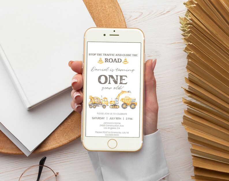 May include: A smartphone displays a construction-themed birthday invitation. The text reads "Stop the traffic and close the road! Daniel is turning ONE year old!" with illustrations of construction vehicles. The invitation includes date, time, and location details.