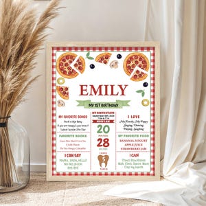 May include: A framed, personalized first birthday milestone poster with a red and white checkered border. The poster features the name "EMILY", pizza illustrations, and details about the child's favorite things, including songs, books, and foods. The poster is 28 inches in height.
