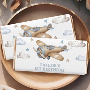 Op de afbeelding: Twee witte chocoladeverpakkingen met een blauw en bruin vliegtuigontwerp. De verpakkingen hebben een blauwe lucht achtergrond met witte wolken en sterren. De tekst op de verpakking luidt "Taylor's 1st Birthday".