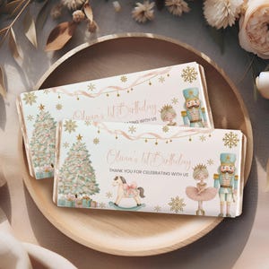 Puede incluir: Dos envoltorios rectangulares de barra de chocolate con temática navideña, árbol decorado, caballito de madera, bailarina y cascanueces. El texto dice Olivia's 1st Birthday, THANK YOU FOR CELEBRATING WITH US, con copos de nieve dorados.