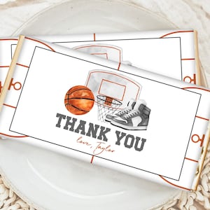 Könnte beinhalten: Zwei weiße Schokoladenriegel-Verpackungen mit Basketball-Motiv. Die Verpackungen zeigen einen Basketball, einen Basketballkorb und Turnschuhe. Der Text "Danke" ist in Grau auf den Verpackungen gedruckt. Der Text "Love, Taylor" ist in Grau unter dem Text "Danke" gedruckt.