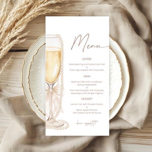 May include: A wedding menu card with a champagne glass illustration and pearl accents. The menu features Entrée, Main, and Dessert options, with the word "Menu" at the top and "bon appétit" at the bottom. The card is on a white plate.