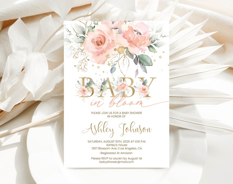 May include: A baby shower invitation with a floral design. The invitation features a watercolor illustration of pink roses and greenery with gold accents. The text "BABY in bloom" is written in gold letters. The invitation includes the details of the baby shower, including the date, time, location, and RSVP information.